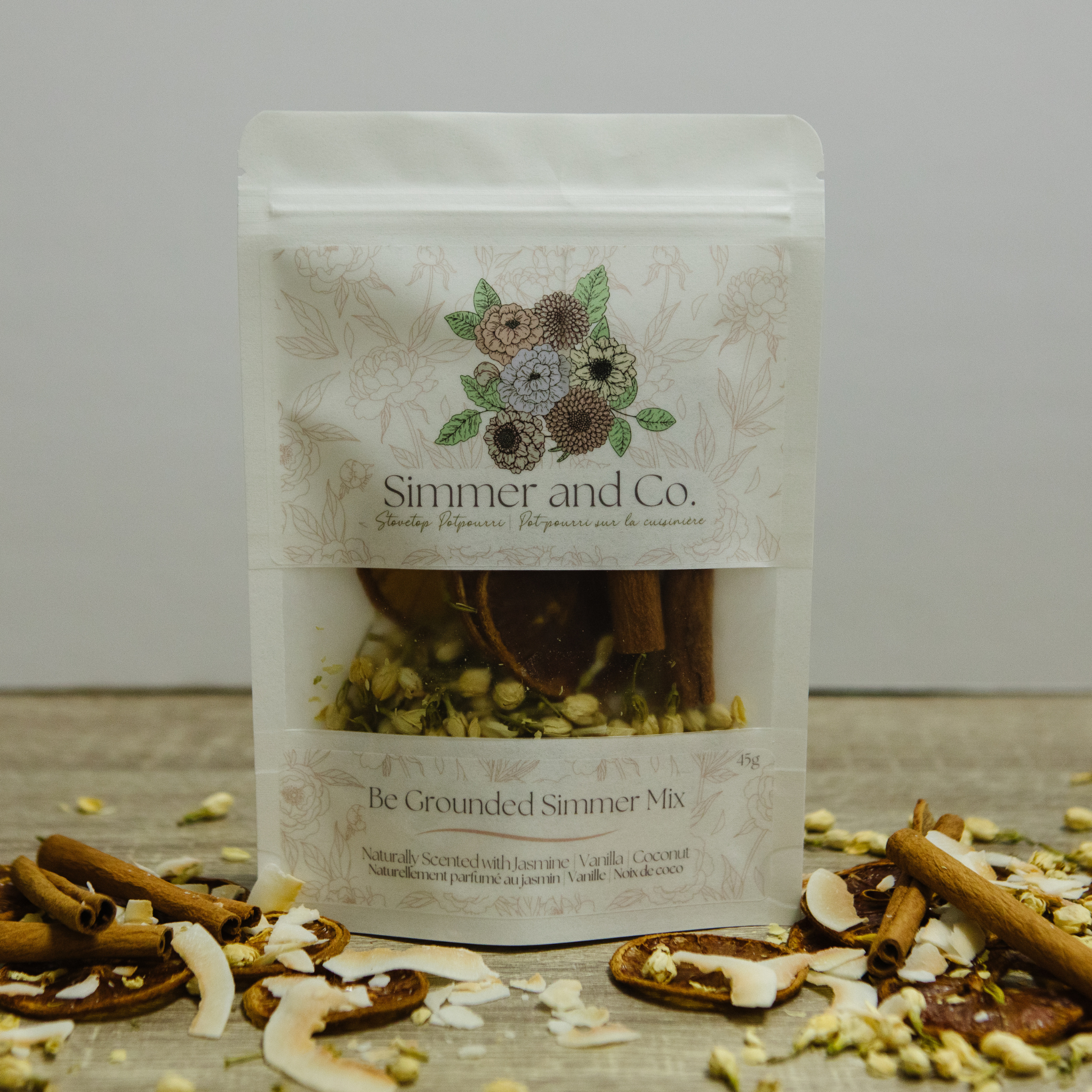Packaged 'Be Grounded Simmer Mix' from Simmer and Co. on a wooden surface with cinnamon sticks and other ingredients.