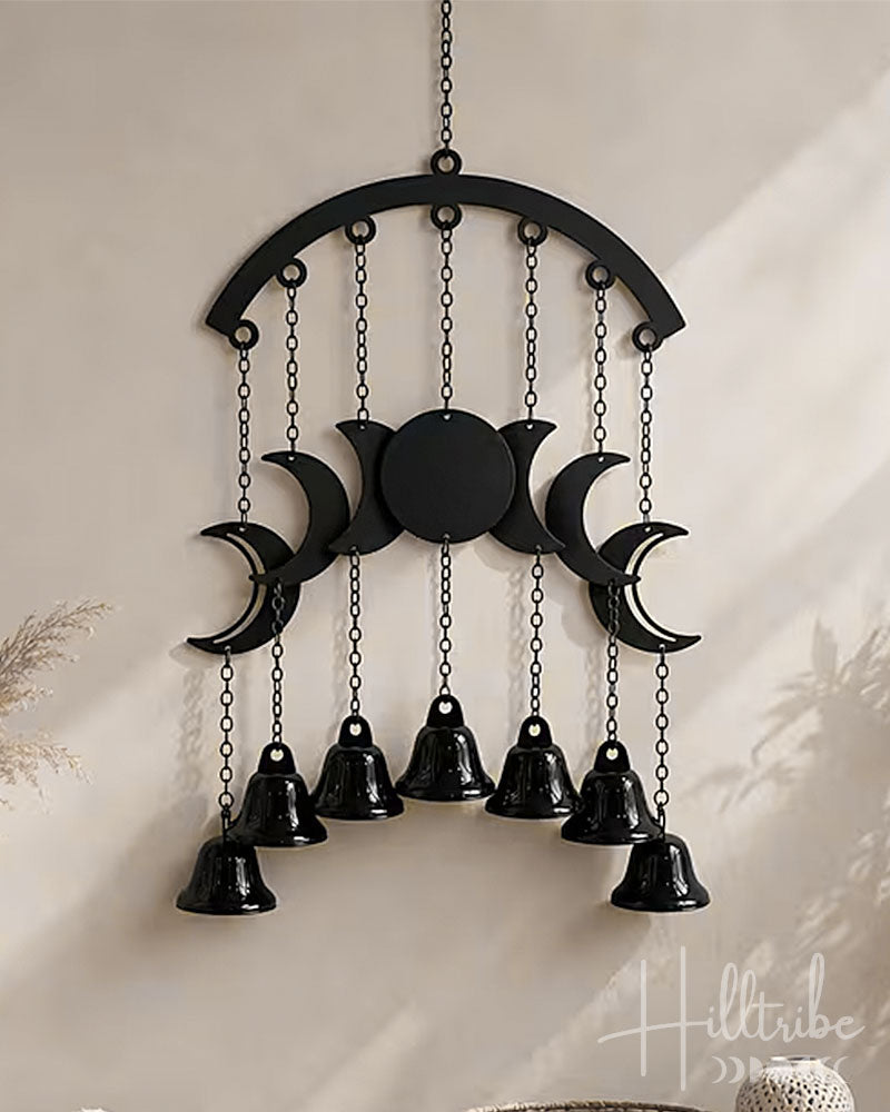 Black metal moon phase wall hanging with seven hanging bells, suspended from a chain against a warm beige wall.