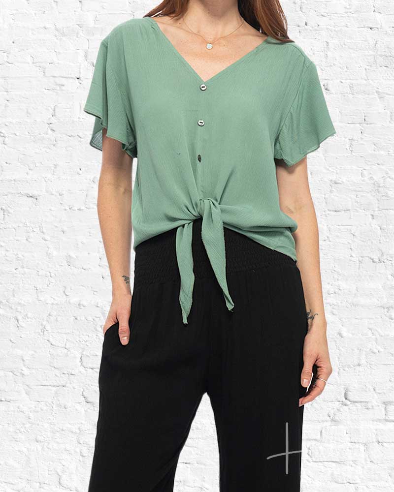 Person wearing a green tied blouse and black pants against a white brick wall.