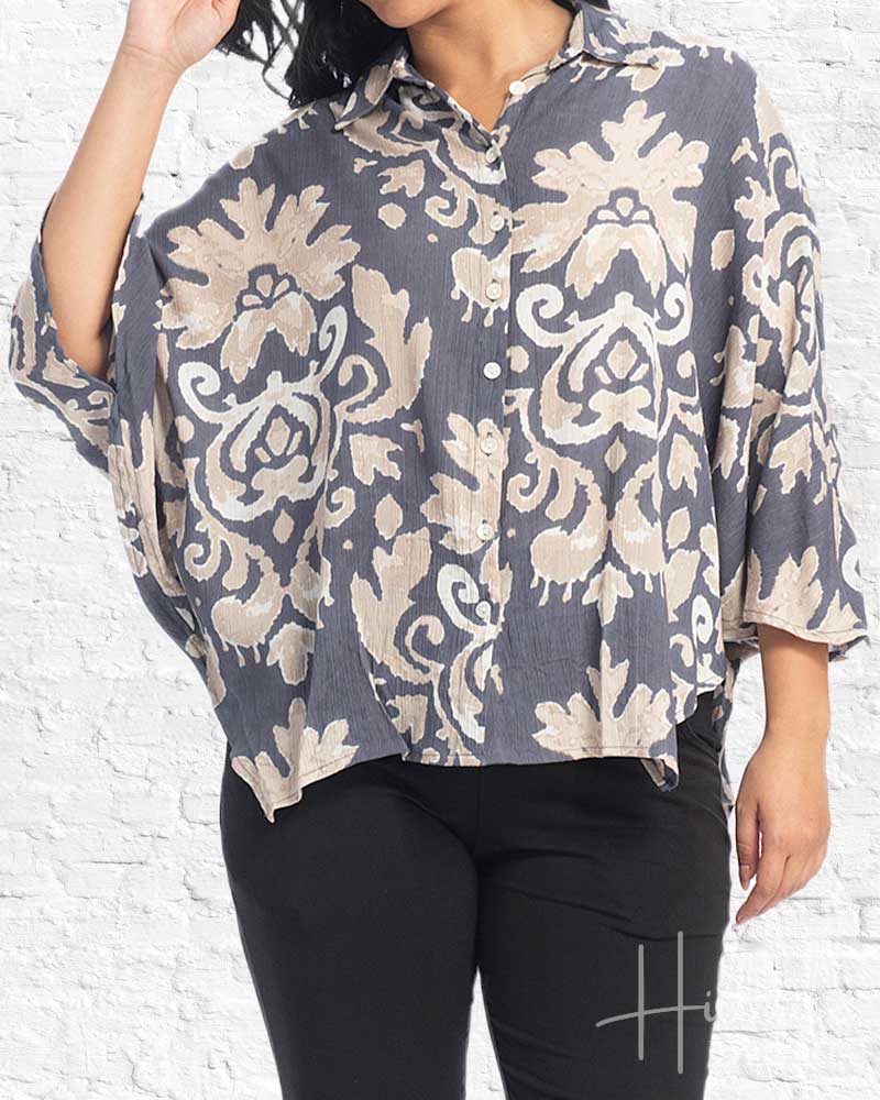A woman wearing a loose, short-sleeve button-up blouse with a gray and beige damask pattern, paired with black pants, standing against a white brick wall.