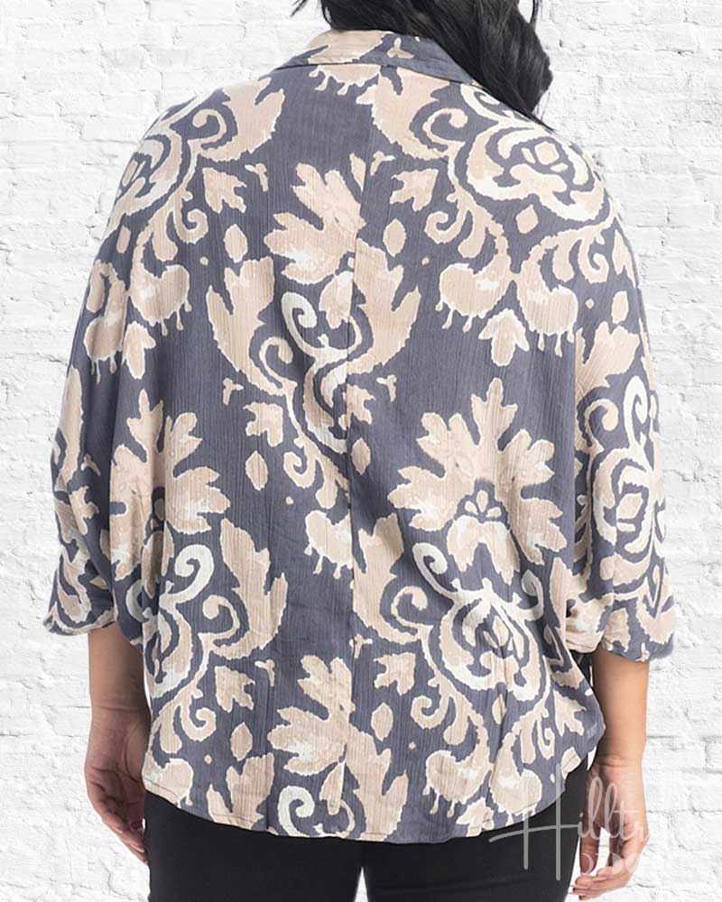 A woman viewed from the back wearing a loose, short-sleeve blouse with a large beige damask-style floral pattern on a muted gray-blue background, paired with black pants against a white brick wall.