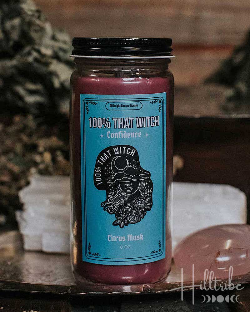 Candle jar with a blue label featuring '100% That Witch' branding on a rustic surface.