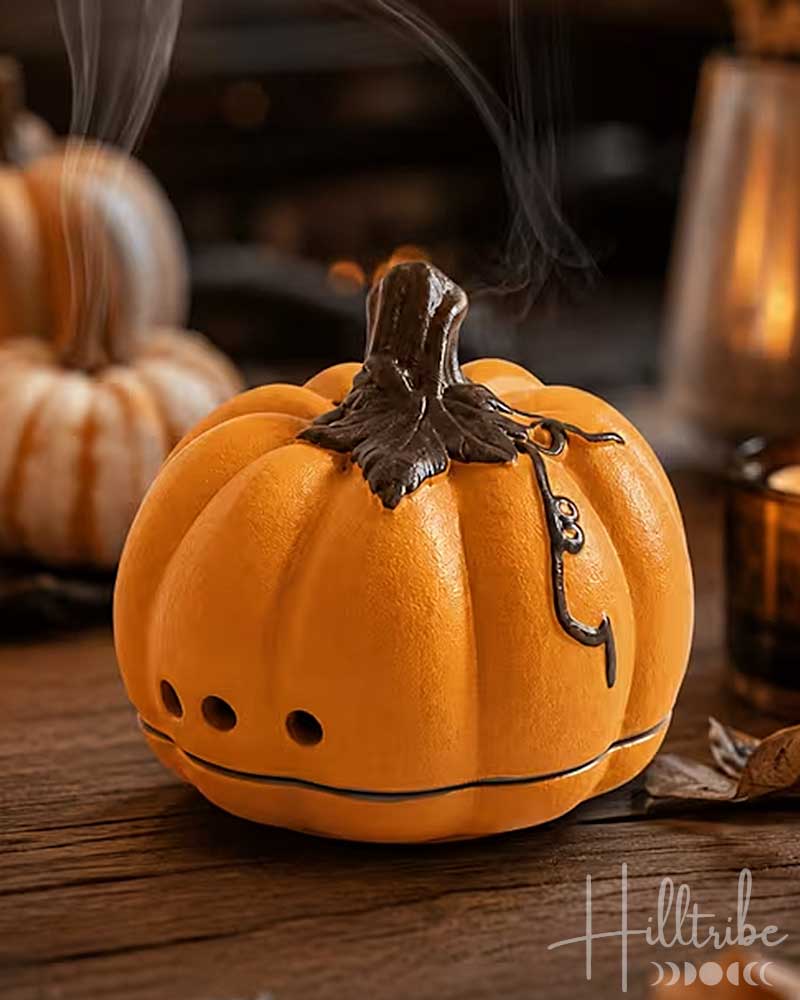 An orange Halloween pumpkin-shaped candle holder with a dark stem, small ventilation holes, and a hanging black spider decoration, shown on a wooden surface with a warm blurred background.