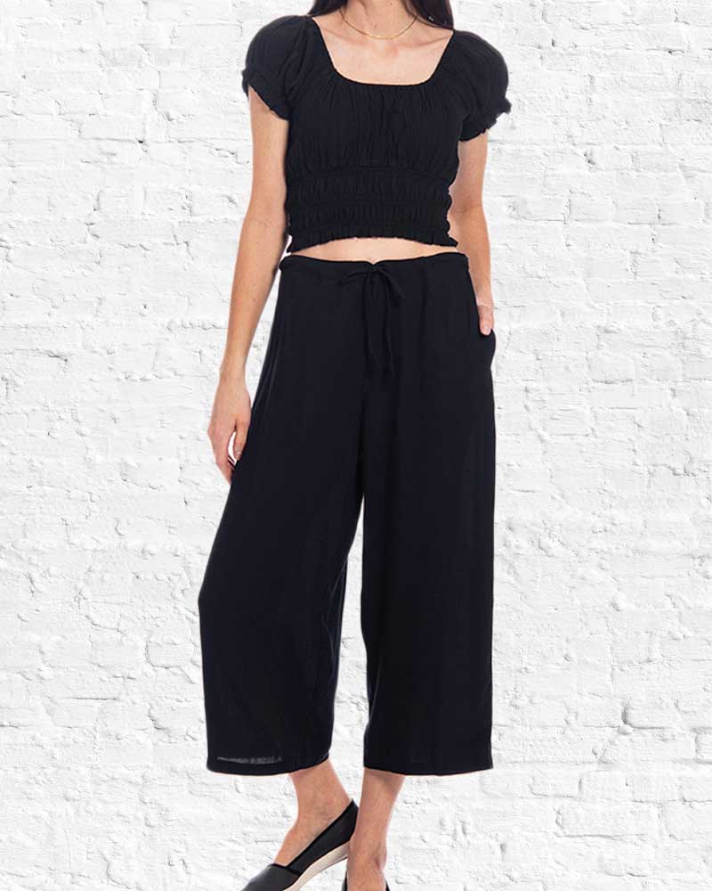 Person wearing a black outfit with a cropped top and wide-leg pants against a white brick wall.