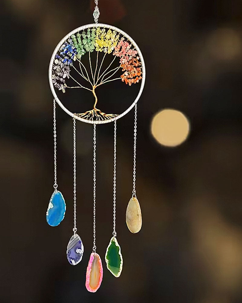 A circular chakra tree wind chime with colorful gemstone accents and five dangling teardrop-shaped crystals on silver chains, set against a dark blurred background with warm bokeh lights.