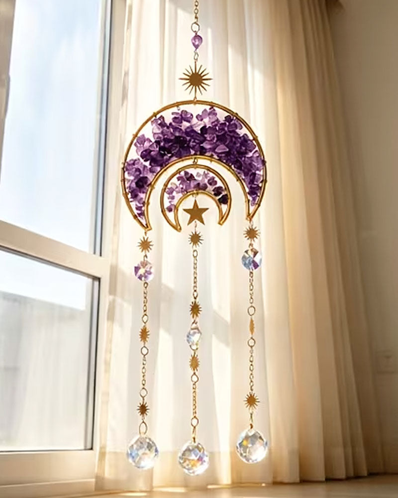A gold crescent moon and star window hanging ornament with purple crystal accents and dangling clear crystal prisms, displayed in front of a window with beige curtains.