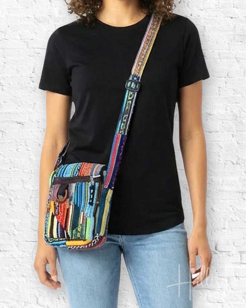 Person wearing a black t-shirt and light blue jeans with a colorful patterned bag over one shoulder against a white brick wall.