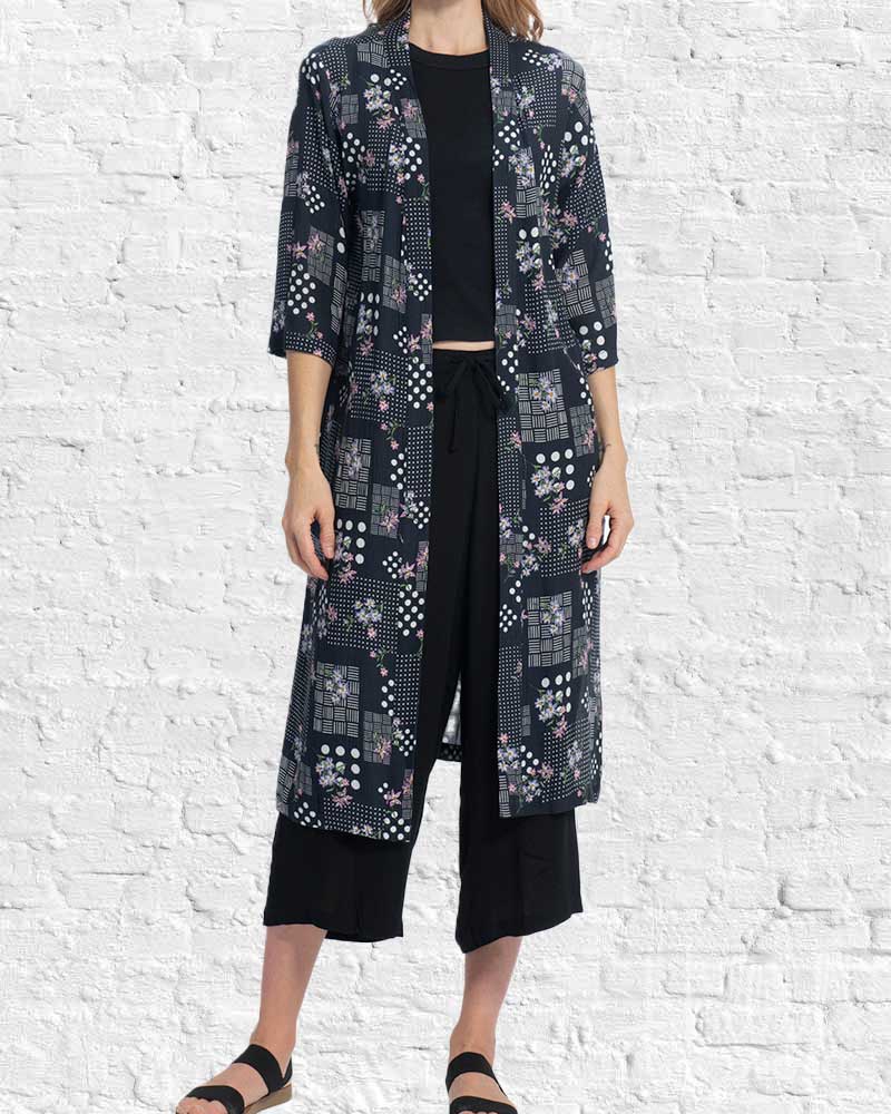 Person wearing a long, dark patterned coat over black attire against a white brick wall.