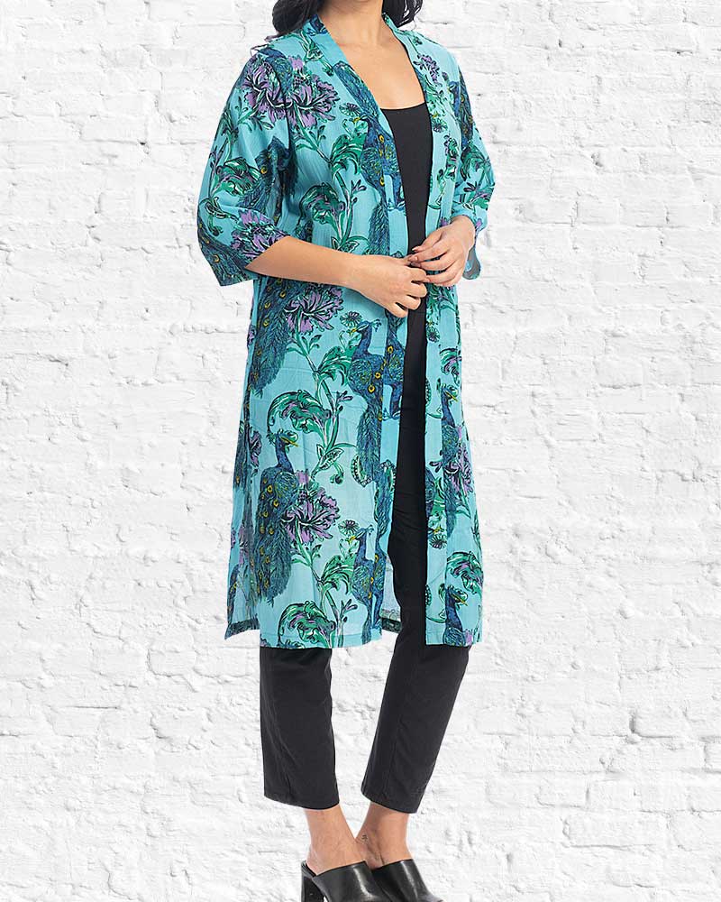 Woman wearing a turquoise floral kimono-style top with black pants against a white brick wall.