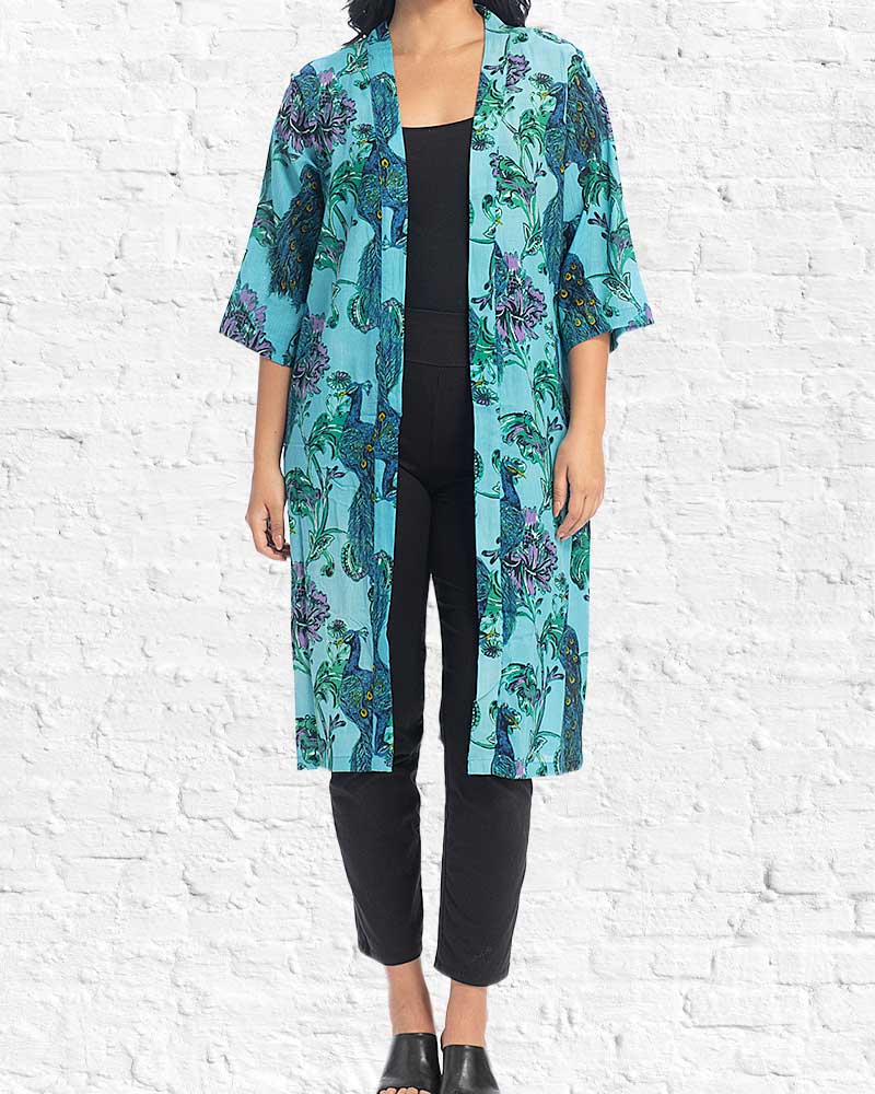Person wearing a turquoise floral kimono over black pants against a white brick wall.