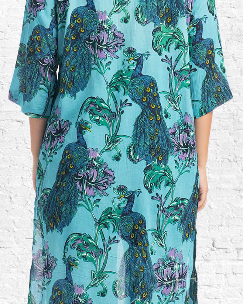 Teal dress with peacock and floral pattern against a white brick wall.