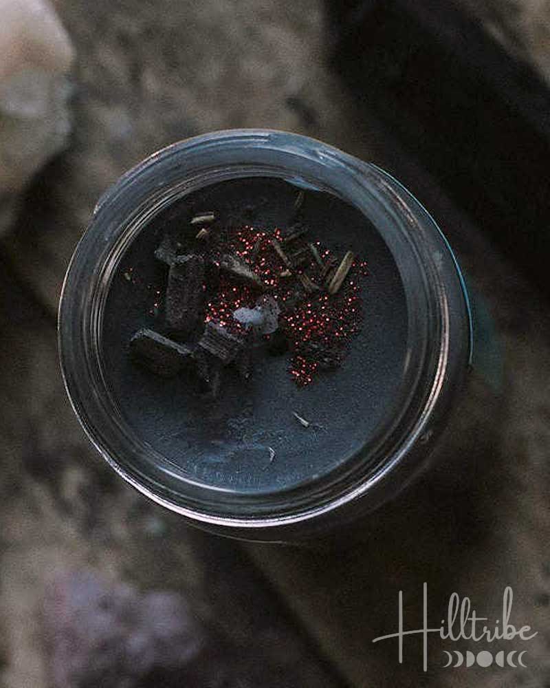 A small glass jar filled with dark gray wax and topped with dried red flowers, styled on a textured stone surface with crystals and a black crystal point.