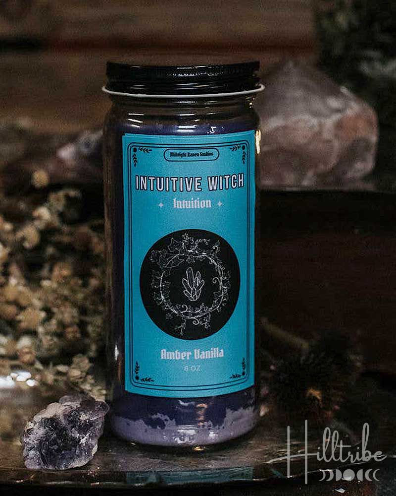 A jar candle labeled 'Intuitive Witch' and 'Berry Vanilla' sits on a rustic metal tray with crystals and dried botanicals against a dark wooden background.