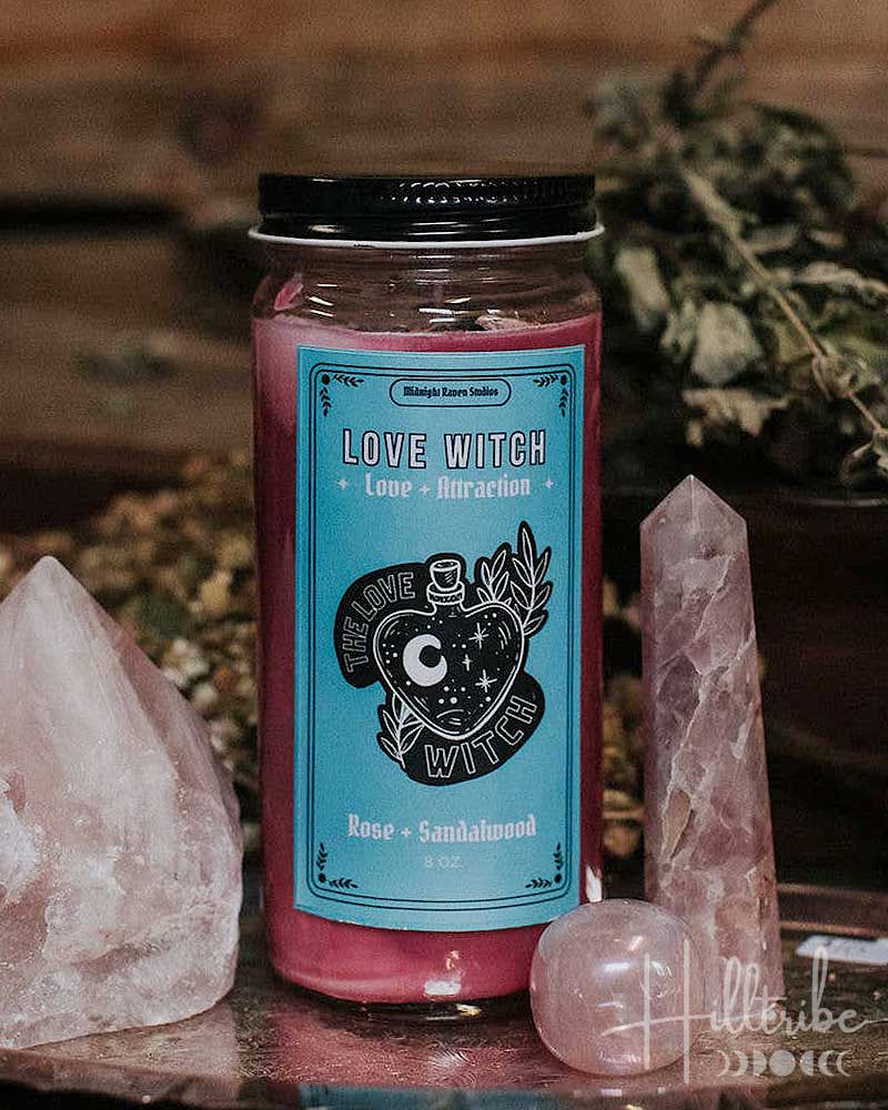 A glass jar candle with a black lid and a turquoise label reading 'Love Witch' and 'Rose + Sandalwood' is styled on a wooden surface with pink crystal points and dried botanicals in the background.