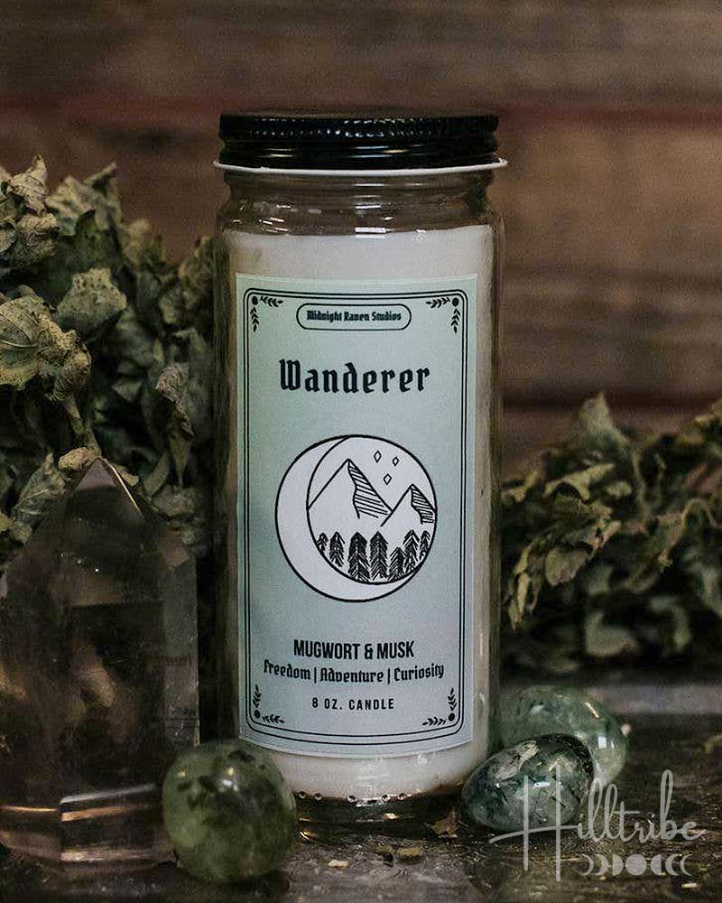 A glass jar candle labelled Wanderer, surrounded by dried botanicals, crystals, and a dark rustic wooden surface against a dark wood plank background.