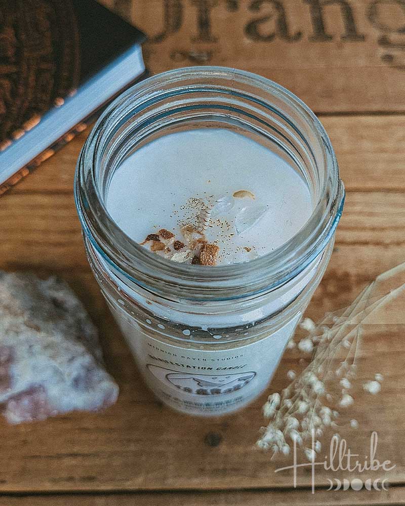 A clear glass jar candle filled with white wax and topped with small dried botanical pieces, placed on a rustic wooden surface next to a crystal and dried flowers.