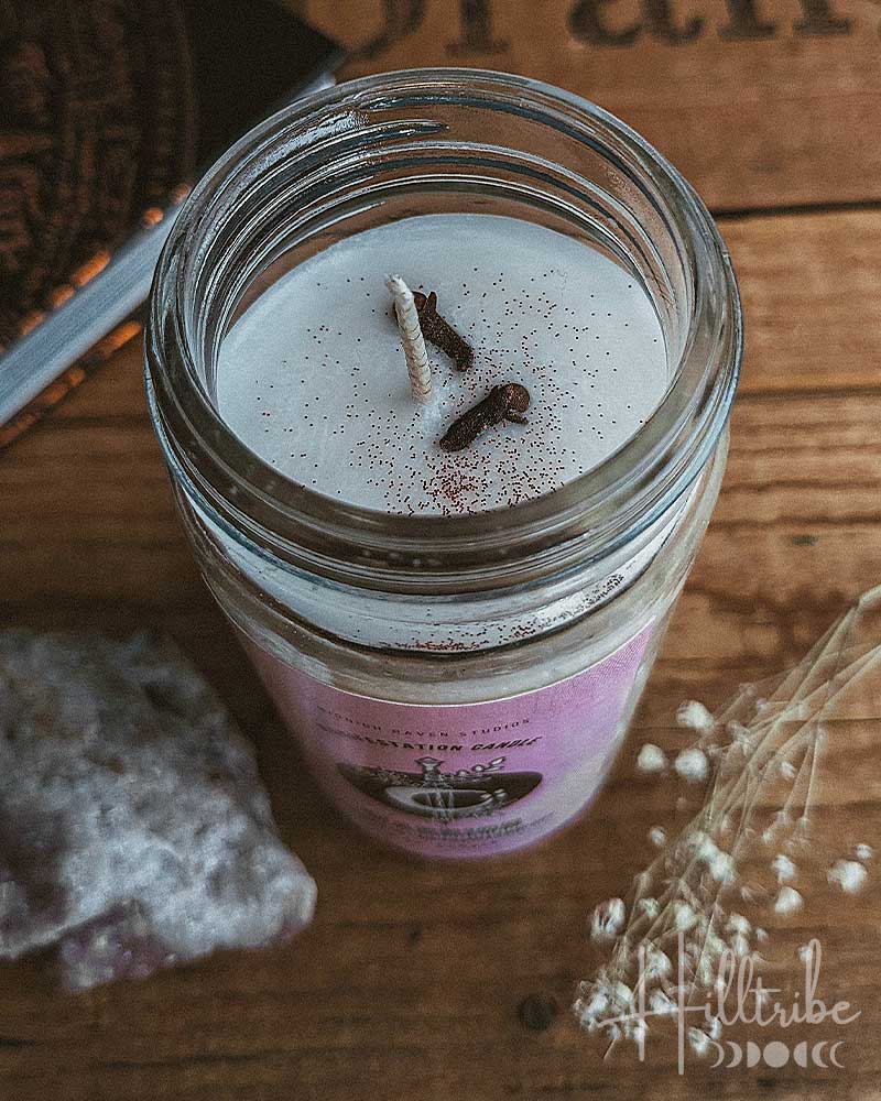A glass jar candle with white wax and one wick, topped with a sprinkle of brown spice, set on a rustic wooden surface next to a purple crystal and dried flowers.