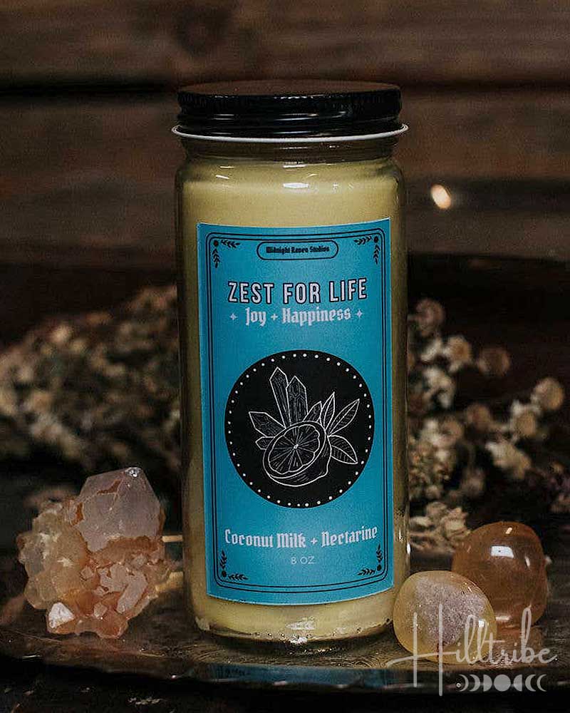 A jar candle labelled 'ZEST FOR LIFE' with a blue label and black lid, placed on a dark tray with crystals and dried flowers against a rustic wooden background.