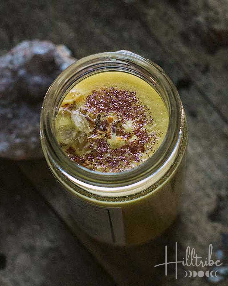 A small clear glass jar filled with a pale yellow balm or wax melt, topped with dried botanicals, resting on a rustic dark wooden surface next to a pink crystal.