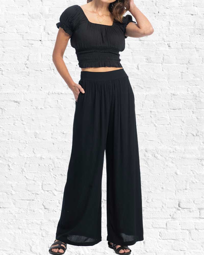 Person wearing a black outfit with a top and wide-leg pants against a white brick wall.