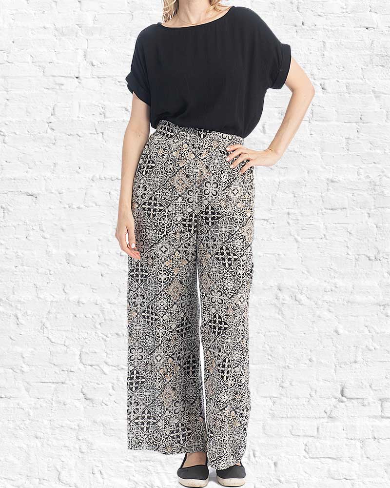Woman wearing a black top and patterned pants against a white brick wall.