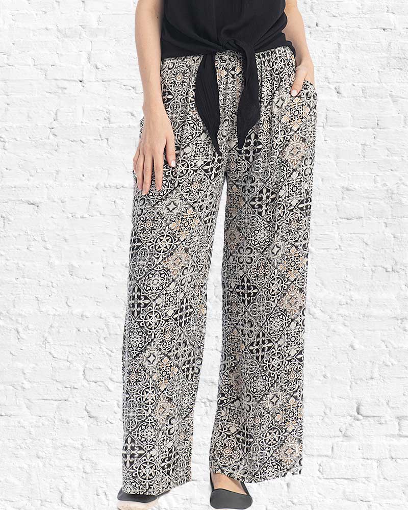 Person wearing black and white patterned pants with a black top against a white brick wall.