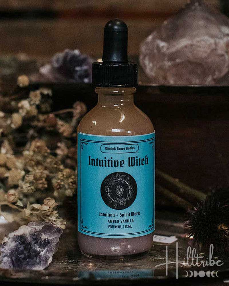 A bottle of Intuitive Witch Sacred Spell Water with a turquoise label and black dropper cap, styled on a rustic wooden surface with crystals and dried botanicals.