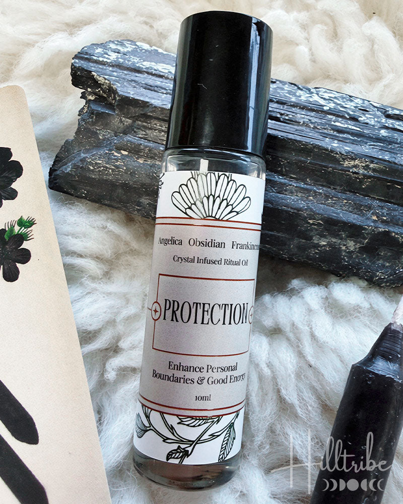A 10ml bottle of Angelica Osmiodan 'Protection' crystal infused ritual oil with a black cap, displayed on a white textured blanket with black wood logs and a decorative card.
