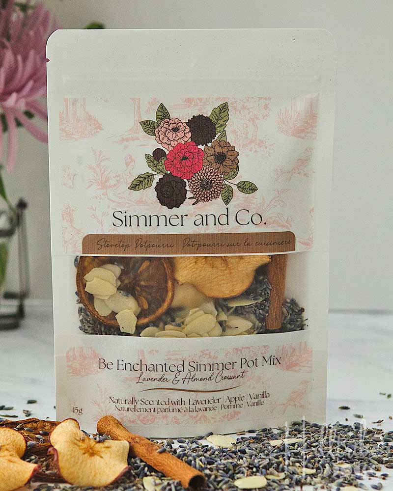 A stand-up pouch of Simmer and Co. Be Enchanted Simmer Pot Mix, shown on a light background with dried apple slices, cinnamon, and loose herbs scattered around the base.