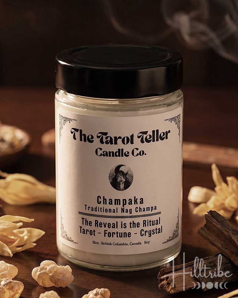 A jar candle labelled 'Champaka' by The Tarot Teller Candle Co. sits on a dark wooden surface with dried flowers, crystals, cinnamon sticks, and smoke in the background.