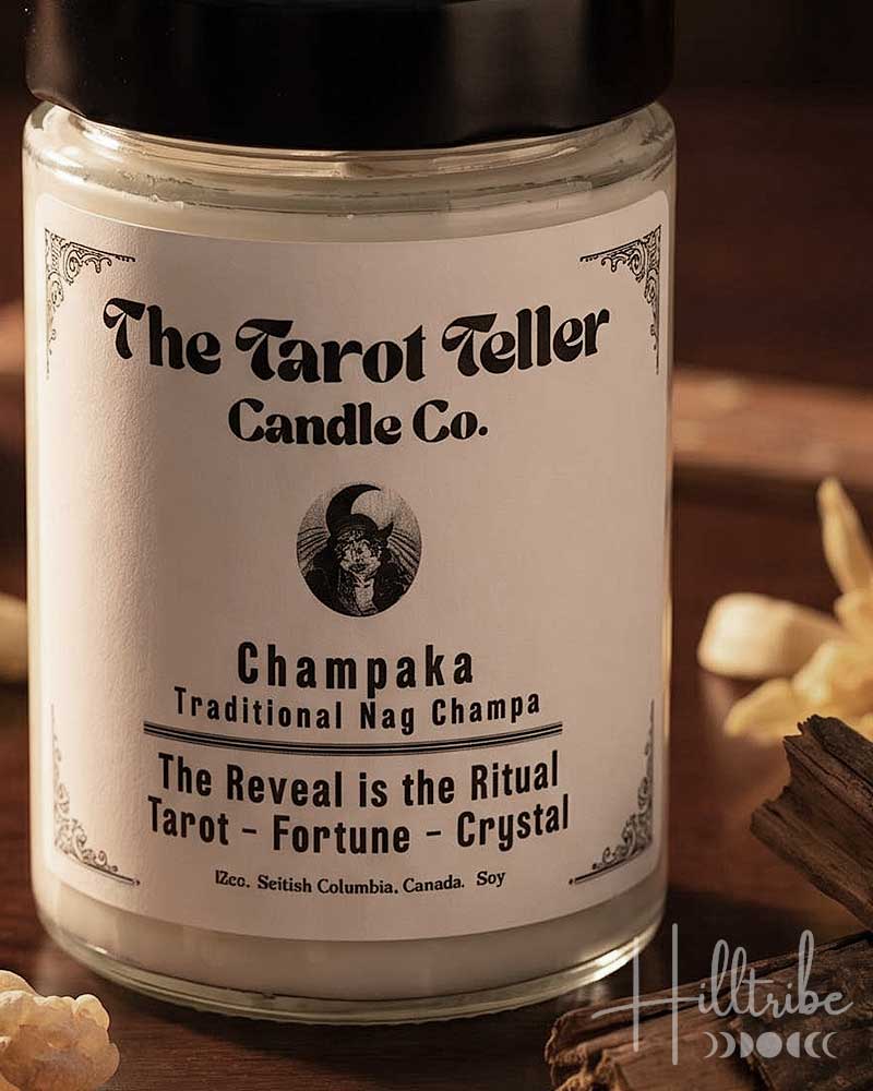 A jar candle labelled 'Champaka' by The Tarot Teller Candle Co. sits on a dark wooden surface with dried flowers, crystals, cinnamon sticks, and smoke in the background.