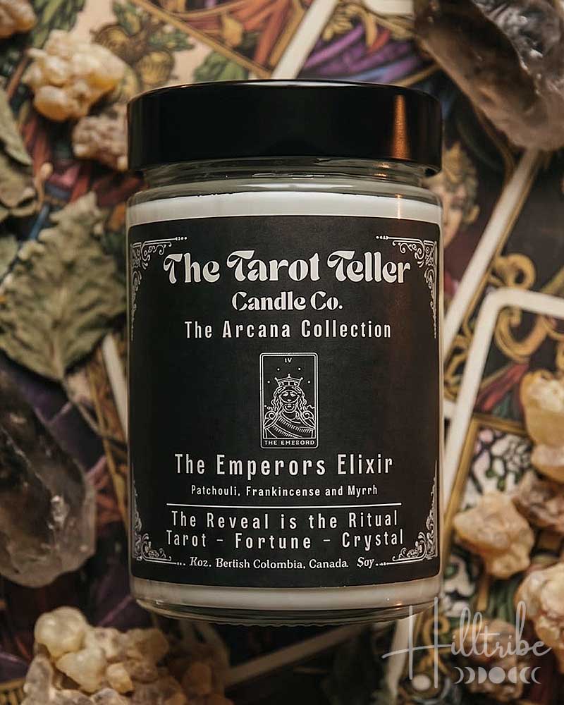 A jar candle labeled 'The Emperors Elixir' by The Tarot Teller Candle Co. is centered over an arranged flat lay of tarot cards, crystals, dried leaves, and wax melts under warm candlelight.