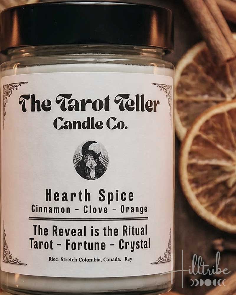 A jar candle labeled 'Hearth Spice' by The Tarot Teller Candle Co. is centered on a dark wooden surface surrounded by dried orange slices, cinnamon sticks, cloves, and crystals.