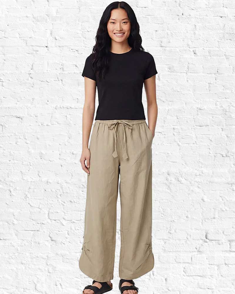Woman wearing a black t-shirt and beige wide-leg pants against a white brick wall.