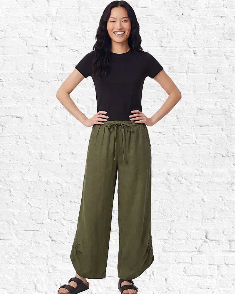 Woman wearing a black t-shirt and green wide-leg pants against a white brick wall.