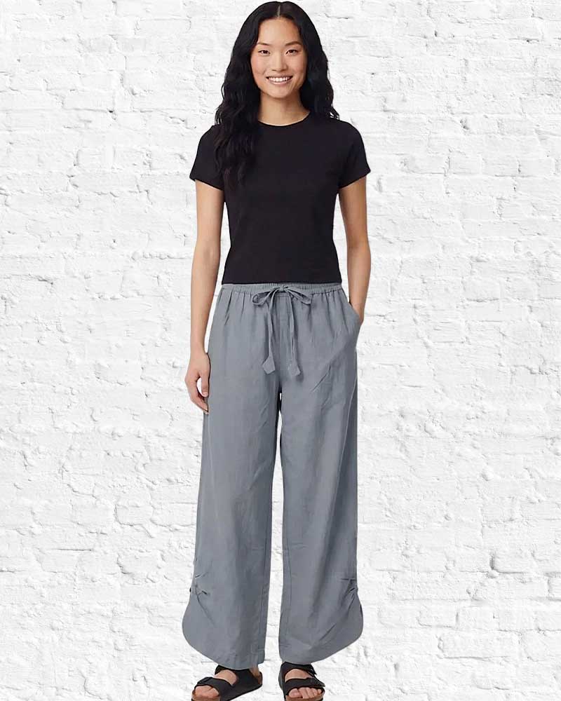 Person wearing a black t-shirt and gray wide-leg pants against a white brick wall.