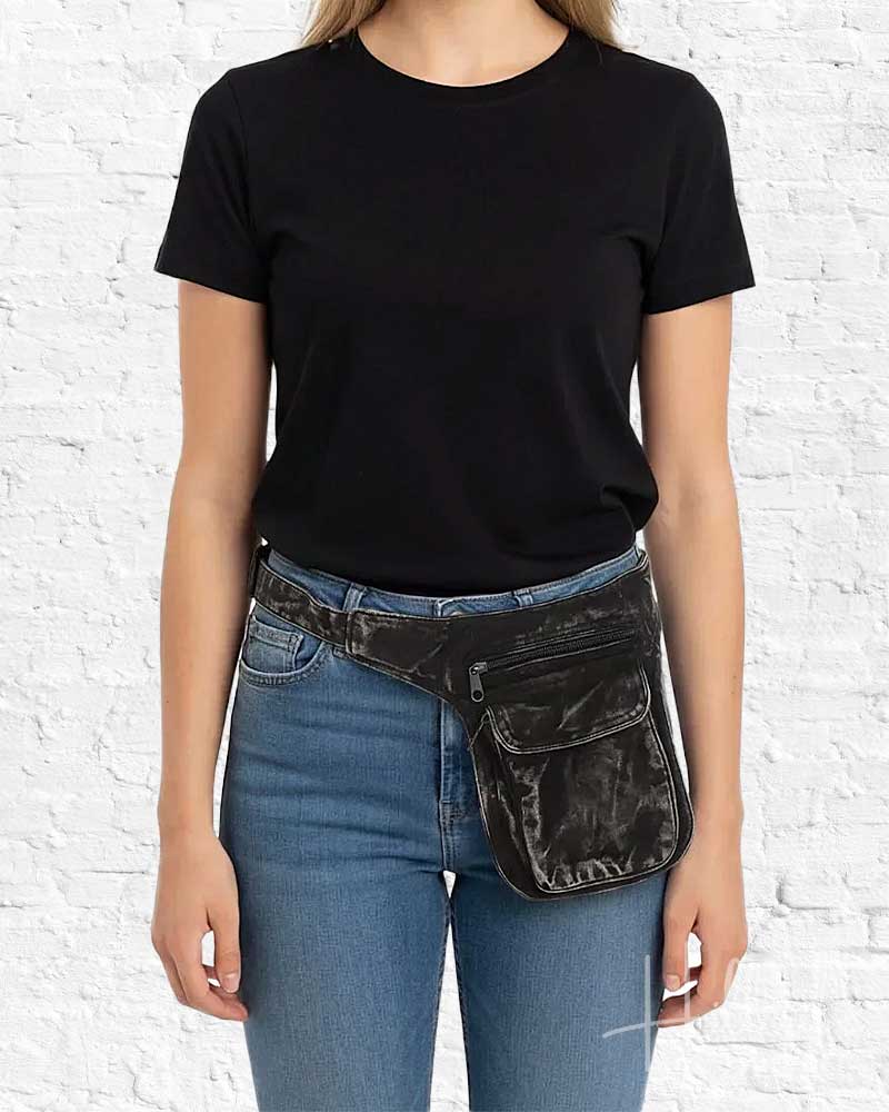 Person wearing a black t-shirt and blue jeans with a black waist bag against a white brick wall.