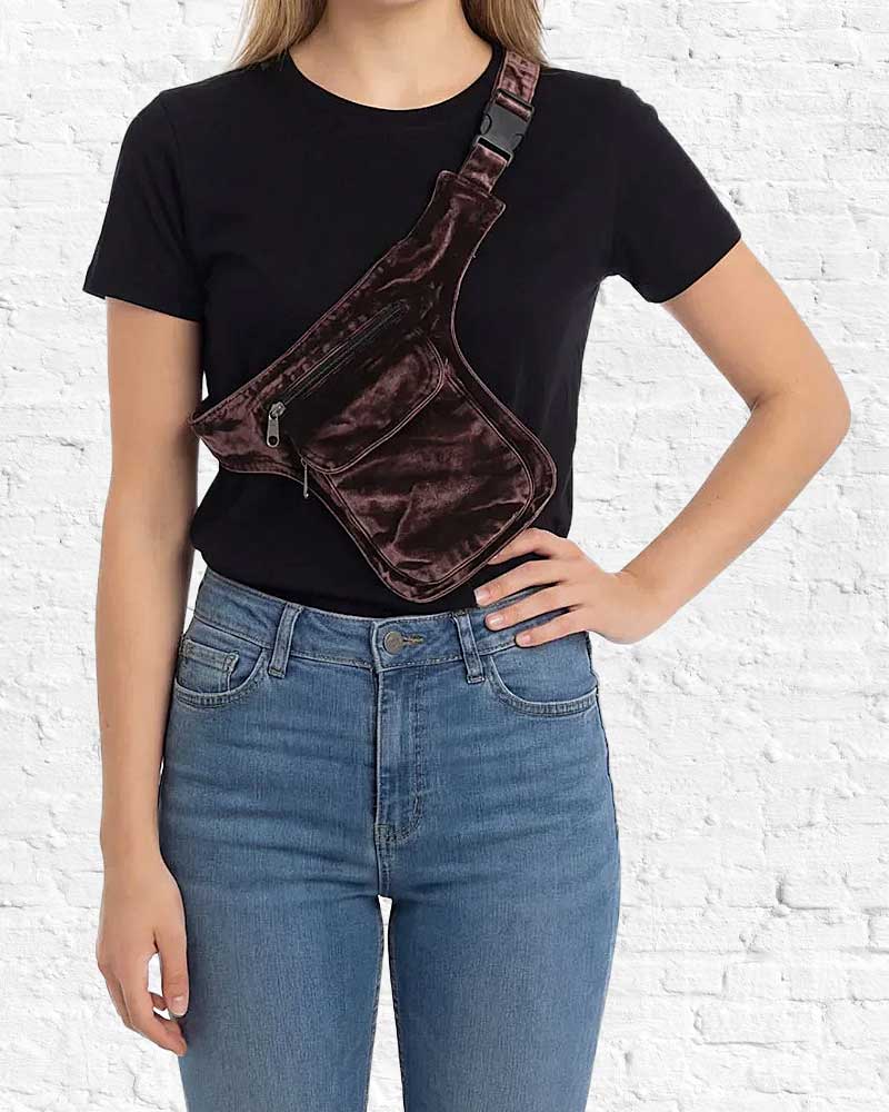 Person wearing a brown velvet fanny pack over a black t-shirt and blue jeans against a white brick wall.