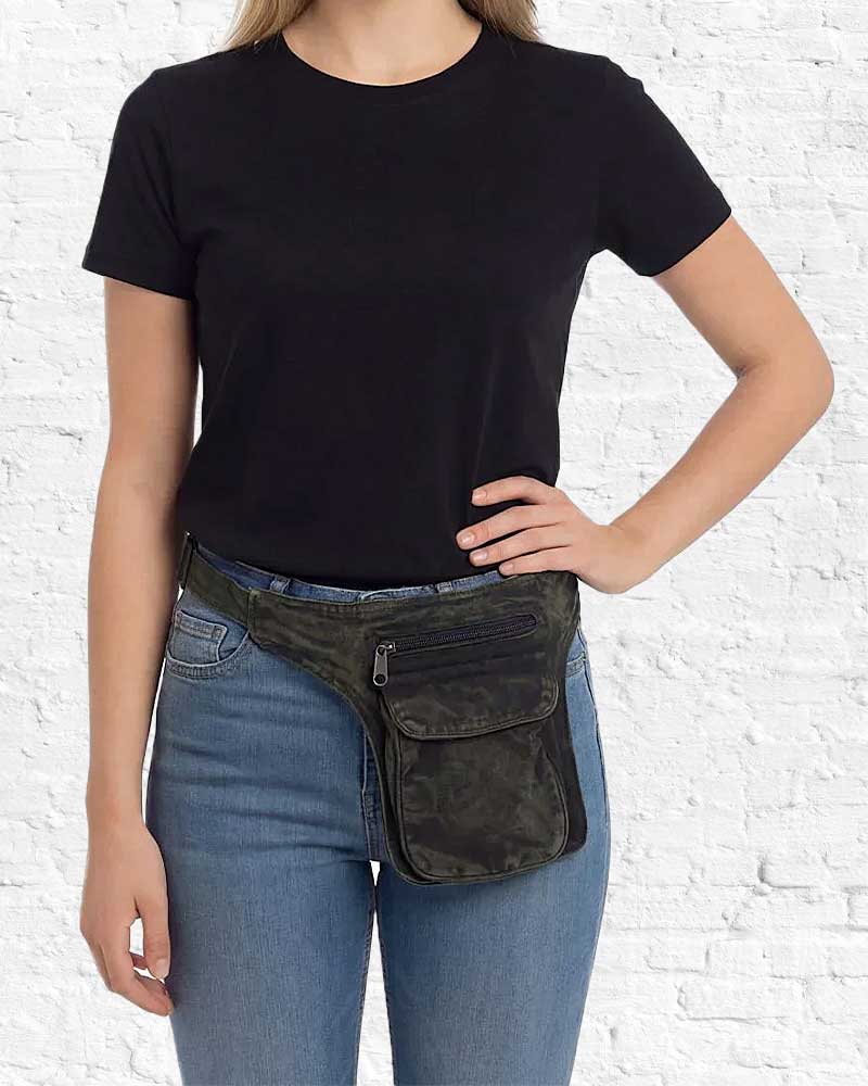 Person wearing a black t-shirt and blue jeans with a dark green waist bag against a white brick wall.