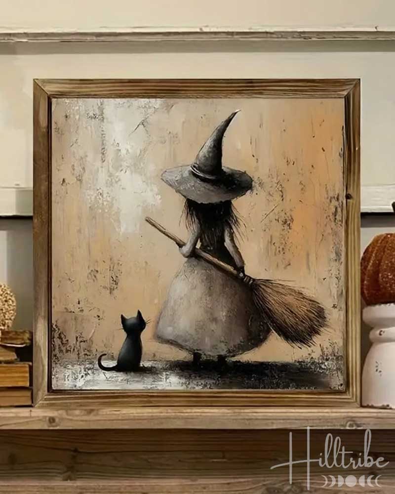 A framed flat wall art print showing a witch in a wide-brimmed hat sweeping a small black cat, displayed on a wooden shelf