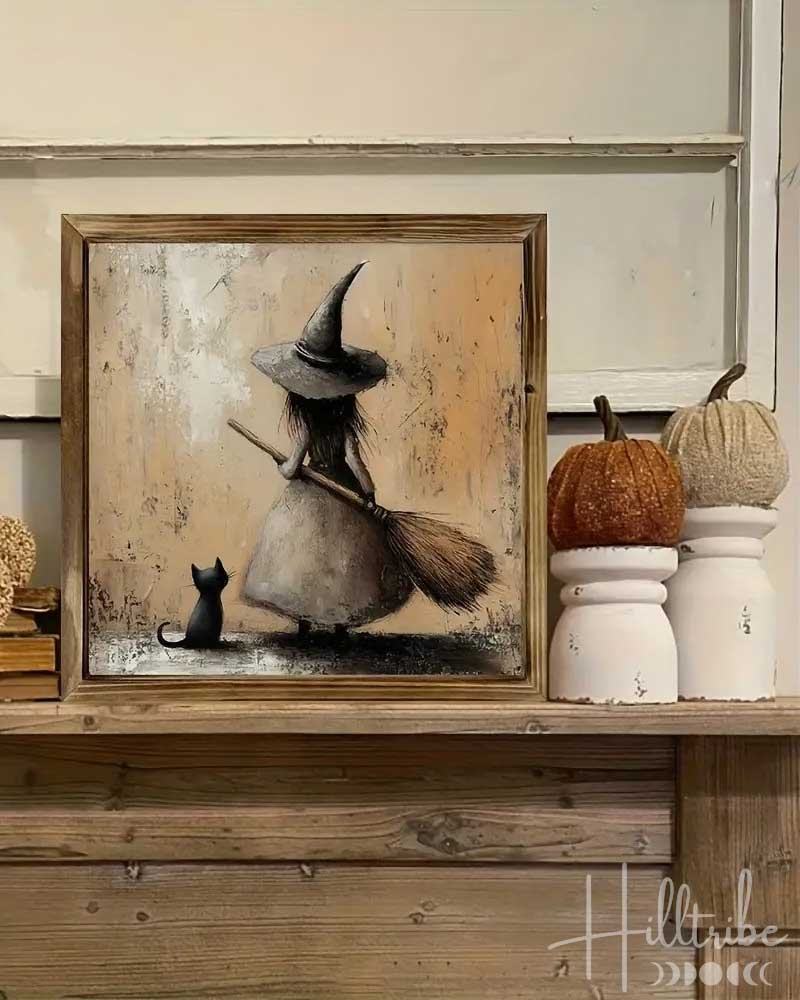 A framed flat wall art print showing a witch in a wide-brimmed hat sweeping a small black cat, displayed on a wooden shelf