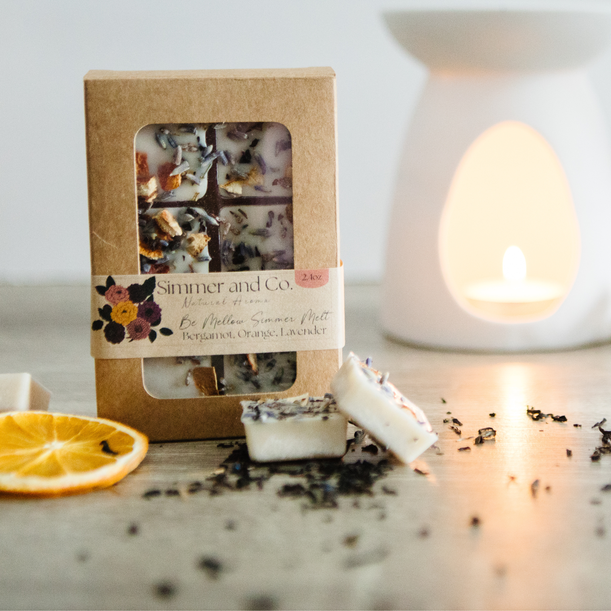 Packaged candles with a decorative box on a surface with a candle and orange slice.