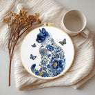 Embroidery hoop with blue floral design and cat silhouette on a textured surface with a cup and dried flowers.