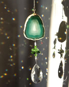 A hanging suncatcher ornament featuring a green agate slice at the top, green faceted beads, and a clear teardrop crystal pendant, shown in bright sunlight against a dark window background with rainbow light leaks.