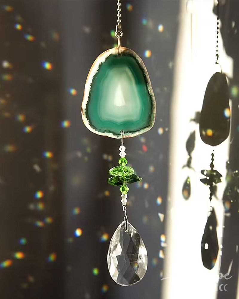 A hanging suncatcher ornament featuring a green agate slice at the top, green faceted beads, and a clear teardrop crystal pendant, shown in bright sunlight against a dark window background with rainbow light leaks.