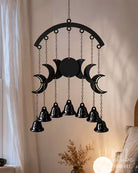 A black metal celestial wall hanging with a triple moon design and nine hanging bells, suspended in a warm, softly lit room.