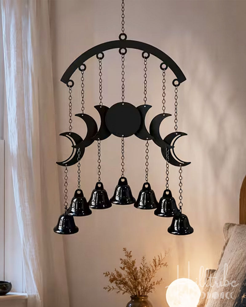 A black metal celestial wall hanging with a triple moon design and nine hanging bells, suspended in a warm, softly lit room.