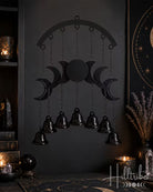 A black metal triple moon and nine bell chandelier hanging on a dark wall in a styled room setting with candles, crystals, and decorative items.