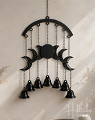 Black metal moon phase wall hanging with seven hanging bells, suspended from a chain against a warm beige wall.