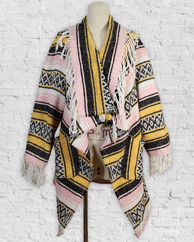 Colorful patterned poncho with fringes on a mannequin against a white brick wall.